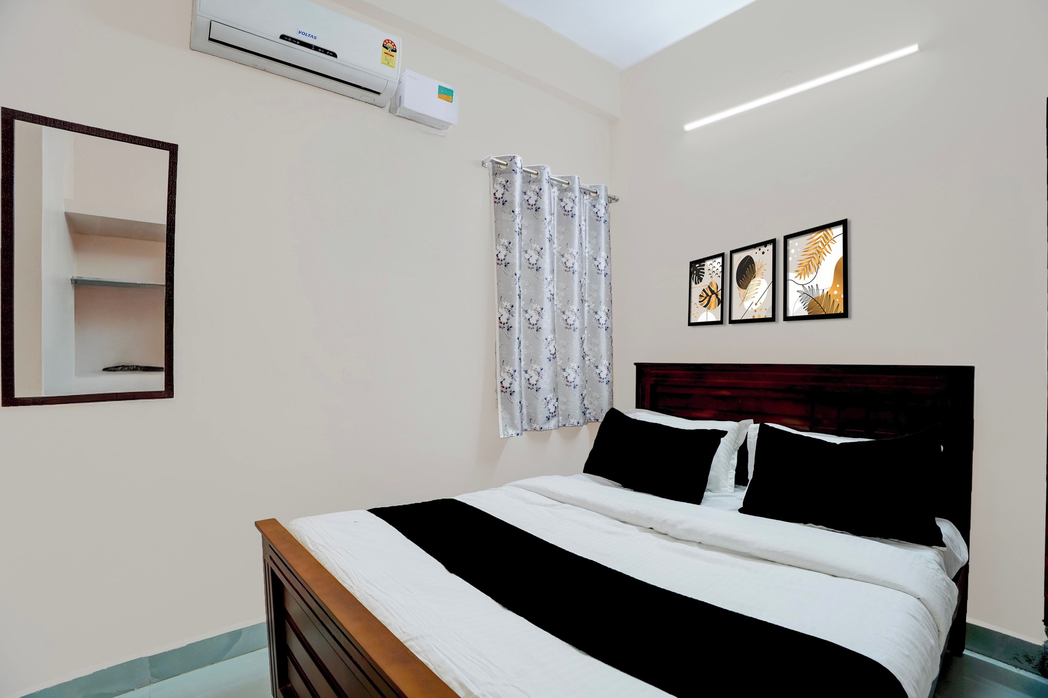 Une chambre à coucher comprenant un lit, un miroir, un appareil de climatisation, une fenêtre avec un rideau à motifs floraux et trois tableaux encadrés accrochés au mur.