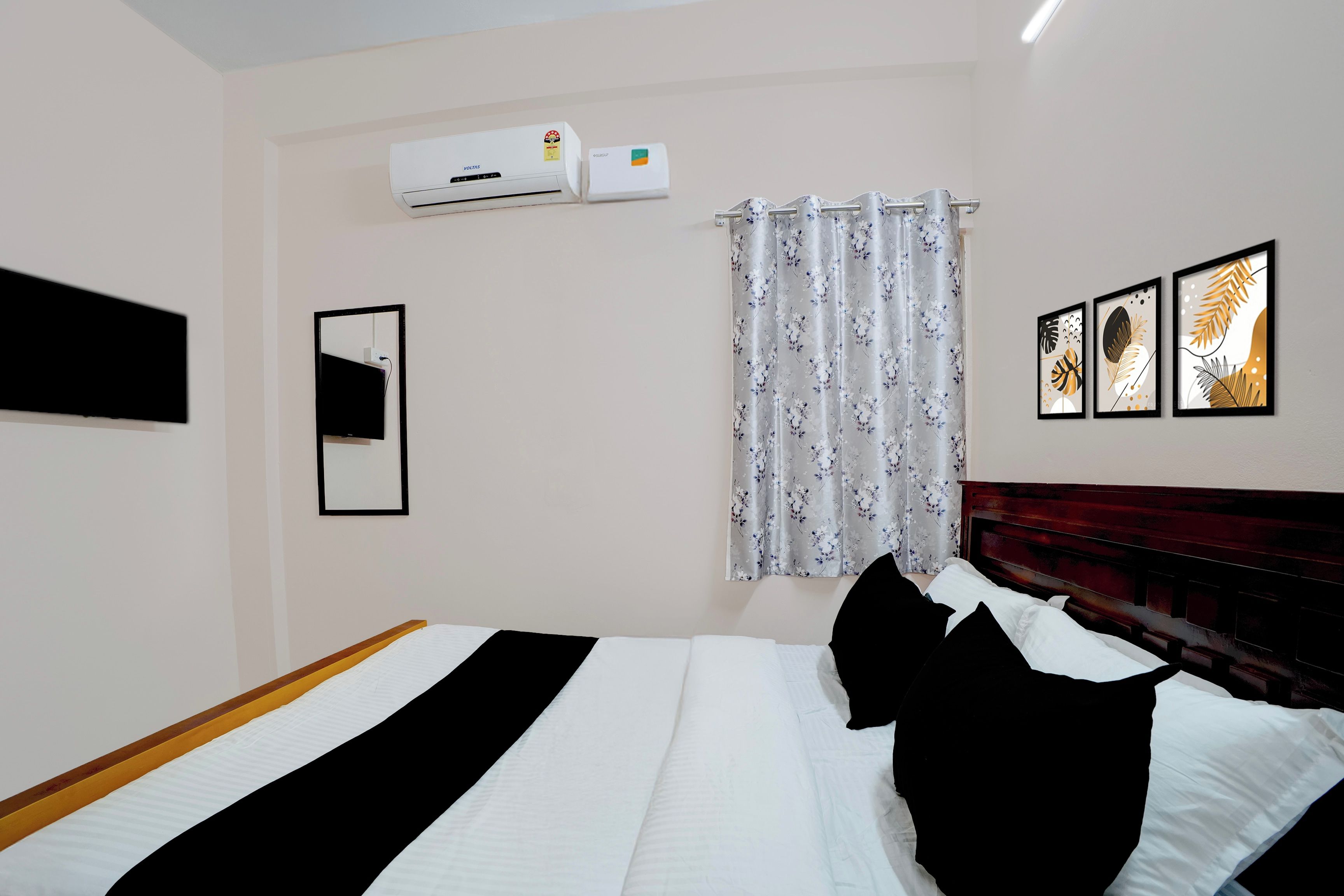 Habitación de hotel con cama, televisor de pantalla plana montado en la pared, aire acondicionado, ventana con cortinas florales y tres cuadros enmarcados.