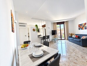A modern living room with a sofa, a dining table set with plates and glasses, and a TV mounted on the wall. - De Vivo Realty - Auralis Home (Massa Lubrense)