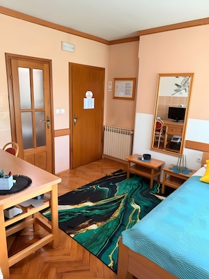 A hotel room with a wooden door, a desk with a chair, a bed with a blue bedspread, a small table with a lamp, and a mirror on the wall. - Apartamento 'Baroko Rooms Apartments' con Wi-Fi y aire acondicionado (Varaždin)