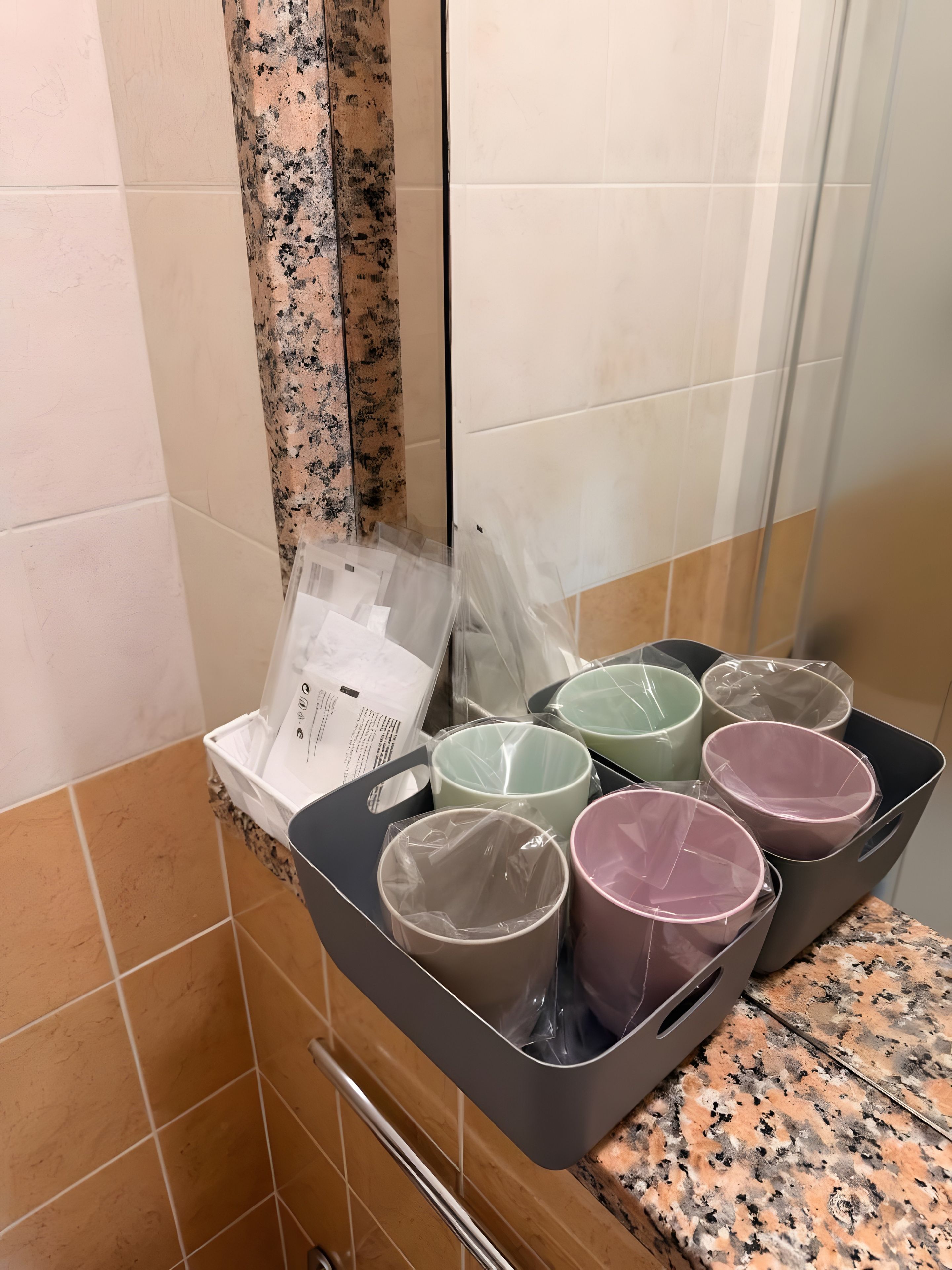 A bathroom with a tiled wall, a shower area, and a countertop with a black tray holding several cups wrapped in plastic.