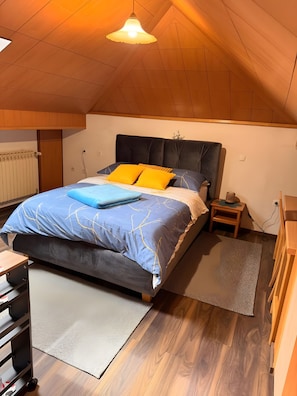 Une chambre avec un plafond en pente, un lit doté d’une tête de lit sombre, une table de chevet et une petite table de nuit.