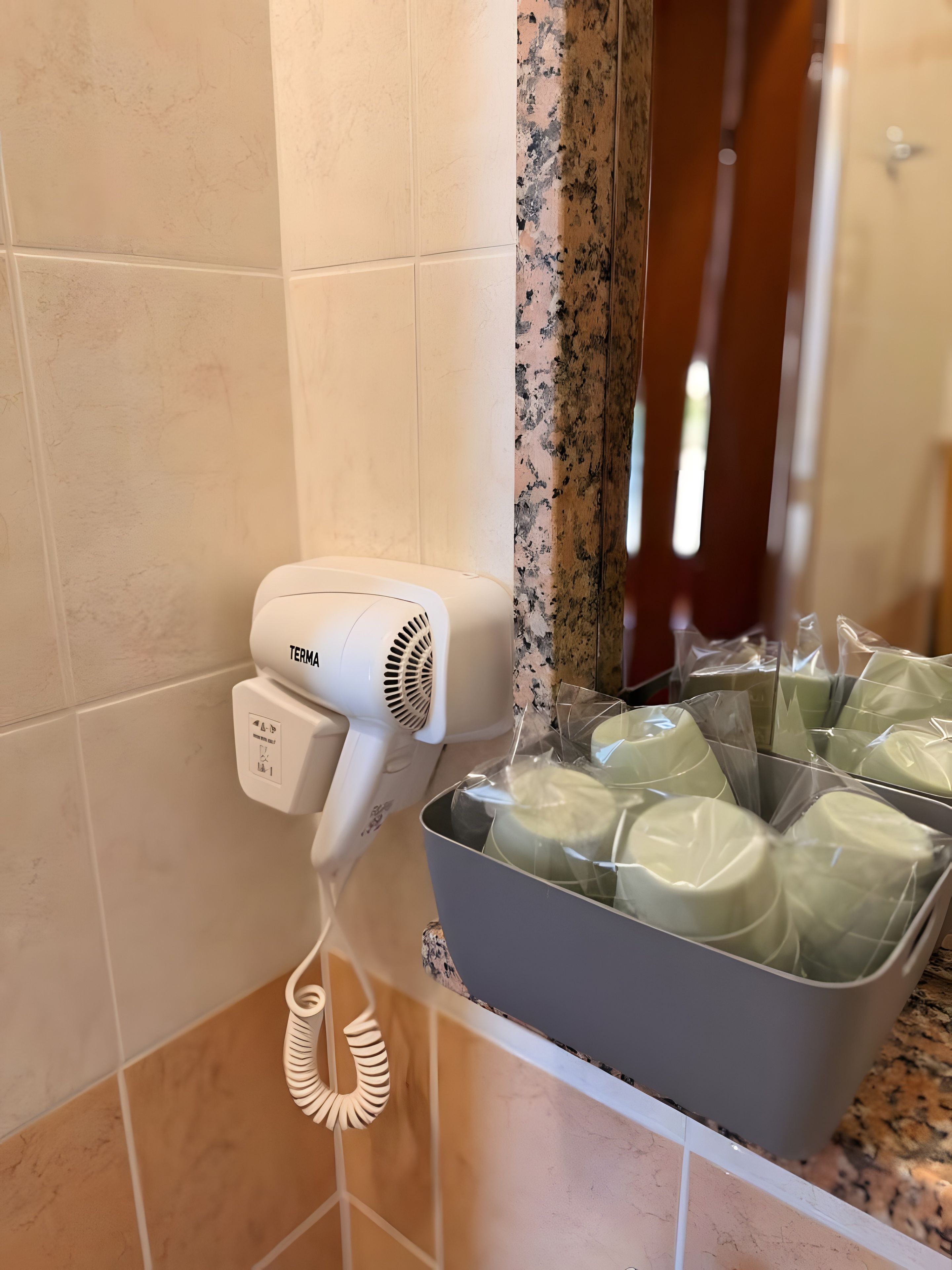 A hairdryer on a wall, a mirror, and a basket of wrapped items.