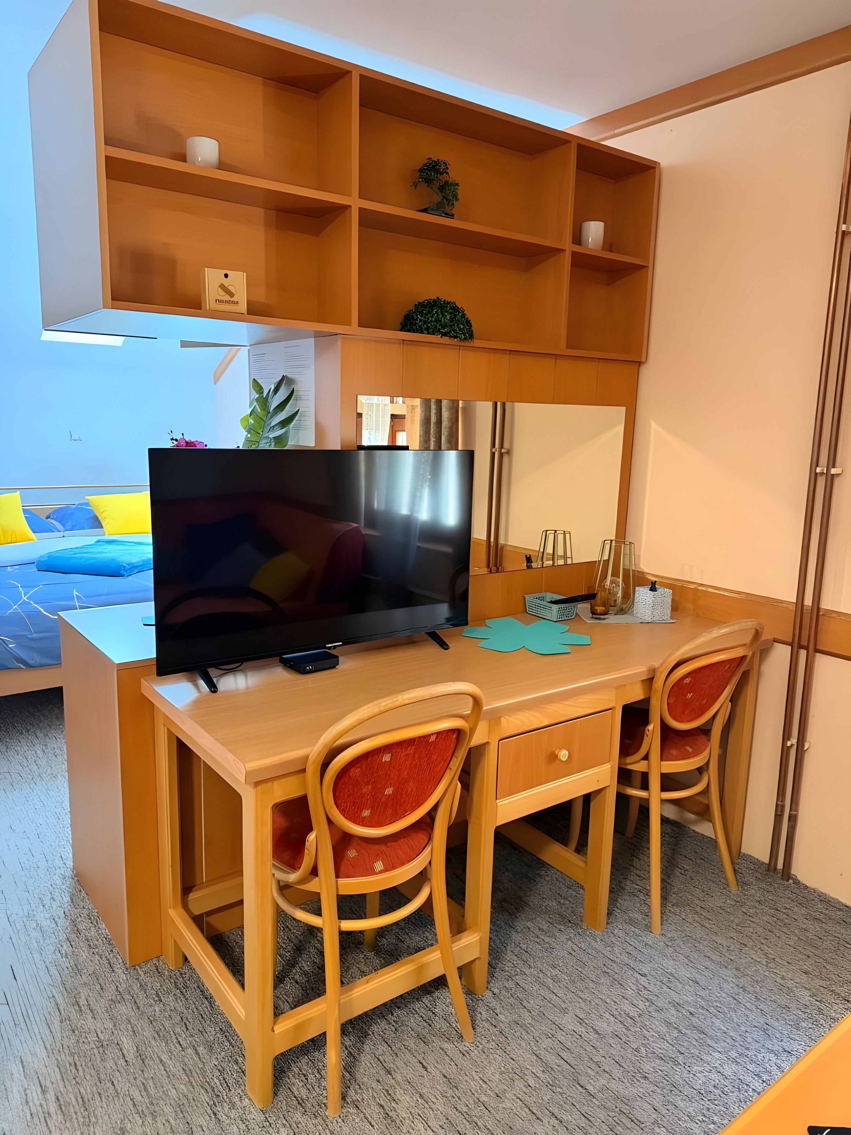 Ett rum med ett skrivbord i trä, två stolar, en tv och en säng med ett blått och gult överkast.