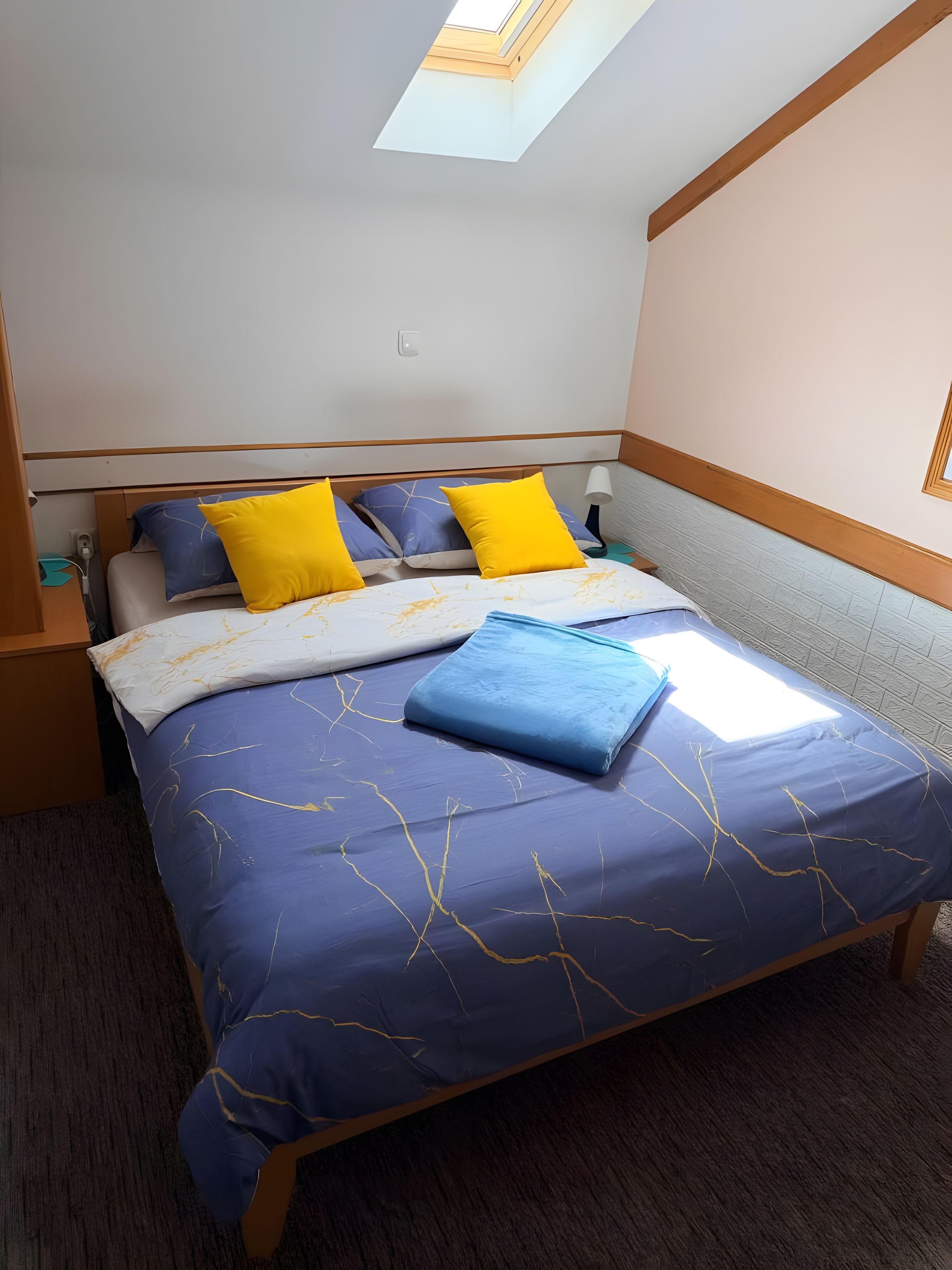 A bedroom with a bed, pillows, a bedside table, a lamp, and a skylight.