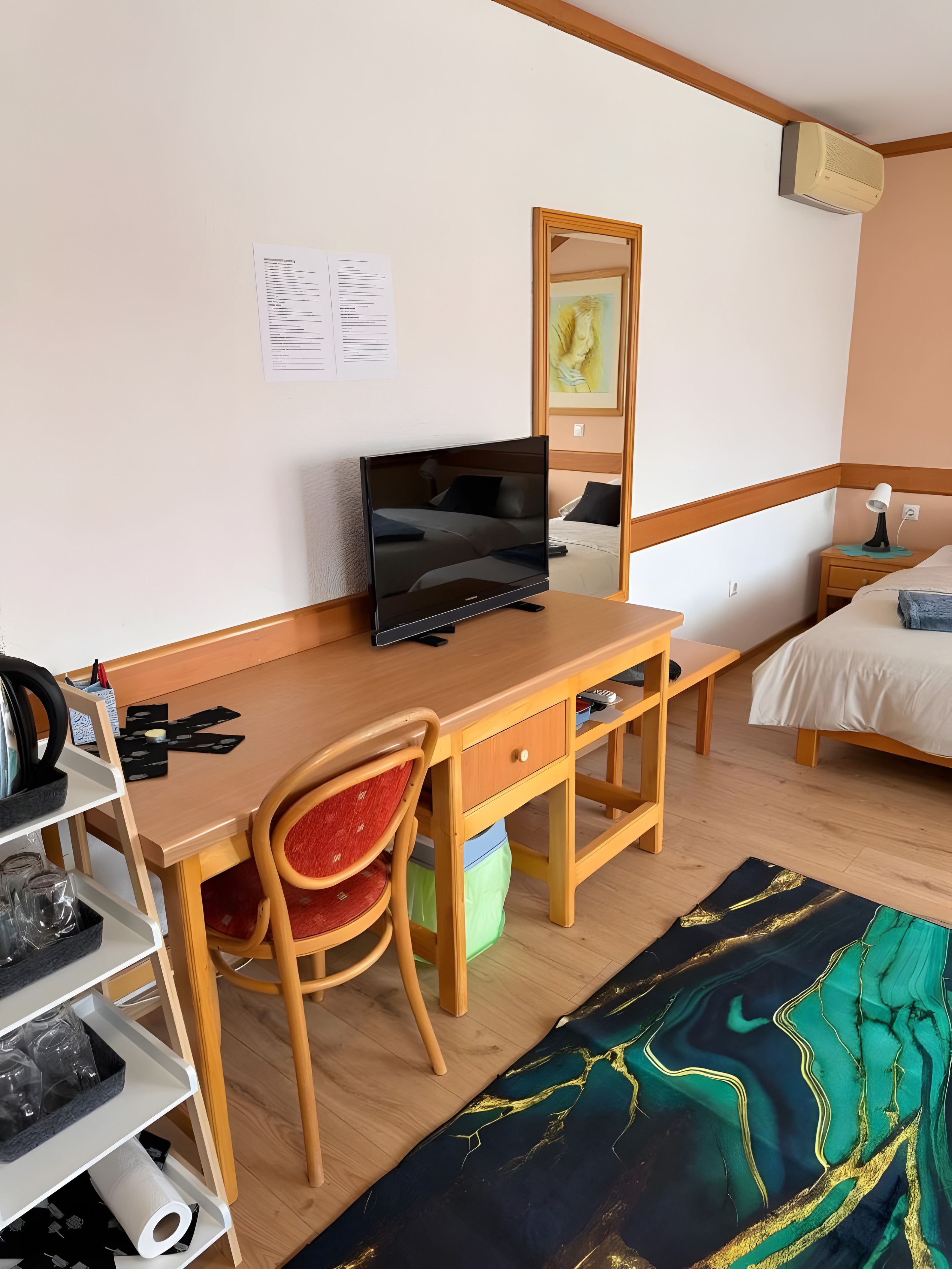 Una habitación con un escritorio de madera, una silla, una cama, una mesita de noche con lámpara, aire acondicionado de pared y un cuadro.