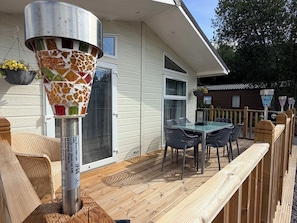 A wooden deck with a table and chairs, a hanging planter with yellow flowers, and a metal pole with a mosaic design. - Boutique Lodge (Exeter)