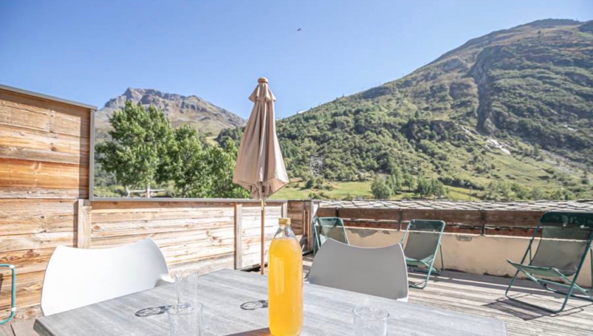 Un'area esterna con terrazza in legno, tavolo, sedie e una bottiglia di succo d'arancia. Si intravede una vista sulle montagne sullo sfondo.