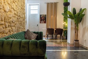 Uma sala de estar com um sofá verde, parede de pedra, uma porta, área de jantar com cadeiras e uma planta em vaso.