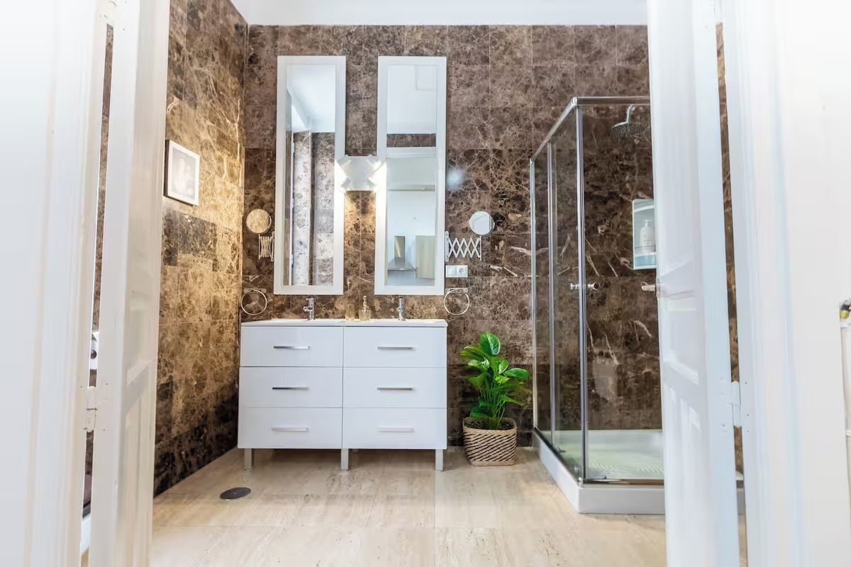 Ein modernes Badezimmer mit Marmorwand, einem weißen Waschtisch, einer Glasdusche und einer Zimmerpflanze.