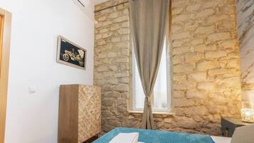 Quarto com parede de pedra, cama com colcha azul, guarda-roupa de madeira e quadro na parede.