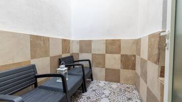 Canto de um quarto com piso de azulejos, um banco e uma pequena mesa com um candeeiro.
