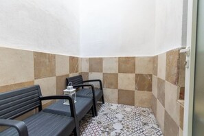 Canto de um quarto com piso de azulejos, um banco e uma pequena mesa com um candeeiro.