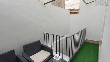 Uma varanda com um pequeno espaço de estar, uma mesa branca e piso de grama sintética verde.