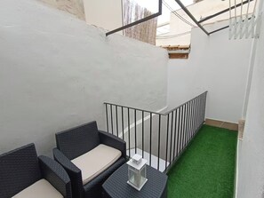 Uma varanda com um pequeno espaço de estar, uma mesa branca e piso de grama sintética verde.