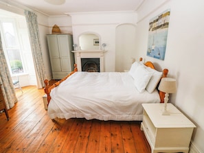 A bedroom with a wooden bed, a white nightstand, a lamp, a fireplace, and a view of the outdoors. - Y Glyn (Borth)