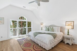 A bedroom with a large bed, a ceiling fan, a window with a view of trees, and a small bedside table with a lamp. - Pier House – Walk to Everything, Worry About Nothing (St Simons Island)