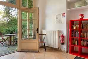 Um quarto com uma grande porta aberta para um jardim, uma cadeira de madeira, uma estante vermelha e um extintor de incêndio na parede.