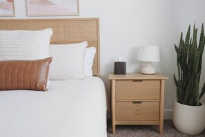 A neatly made bed with white bedding and a brown pillow, a bedside table with a lamp and a tissue box, and a large potted plant. - Bright 2-Bedroom Home with Mountain Views (Hyrum)