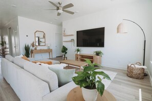 A modern living room with a sofa, coffee table, and TV. There is a ceiling fan, a floor lamp, and a basket. - Bright 2-Bedroom Home with Mountain Views (Hyrum)