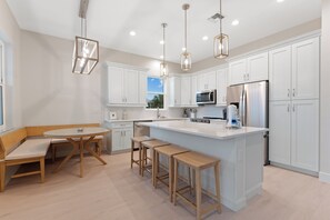 A modern kitchen with white cabinetry, a central island, bar stools, pendant lighting, and a view of the outdoors through a window. - 🌴Sandpiper Shores | Heated Pool + Steps to Beach 🌴 (Fort Myers Beach)