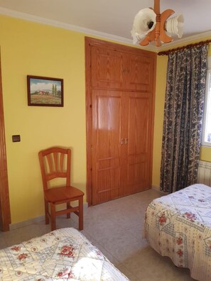 A bedroom with a wooden wardrobe, a chair, two beds with floral bedding, a ceiling fan, and a window with curtains.
