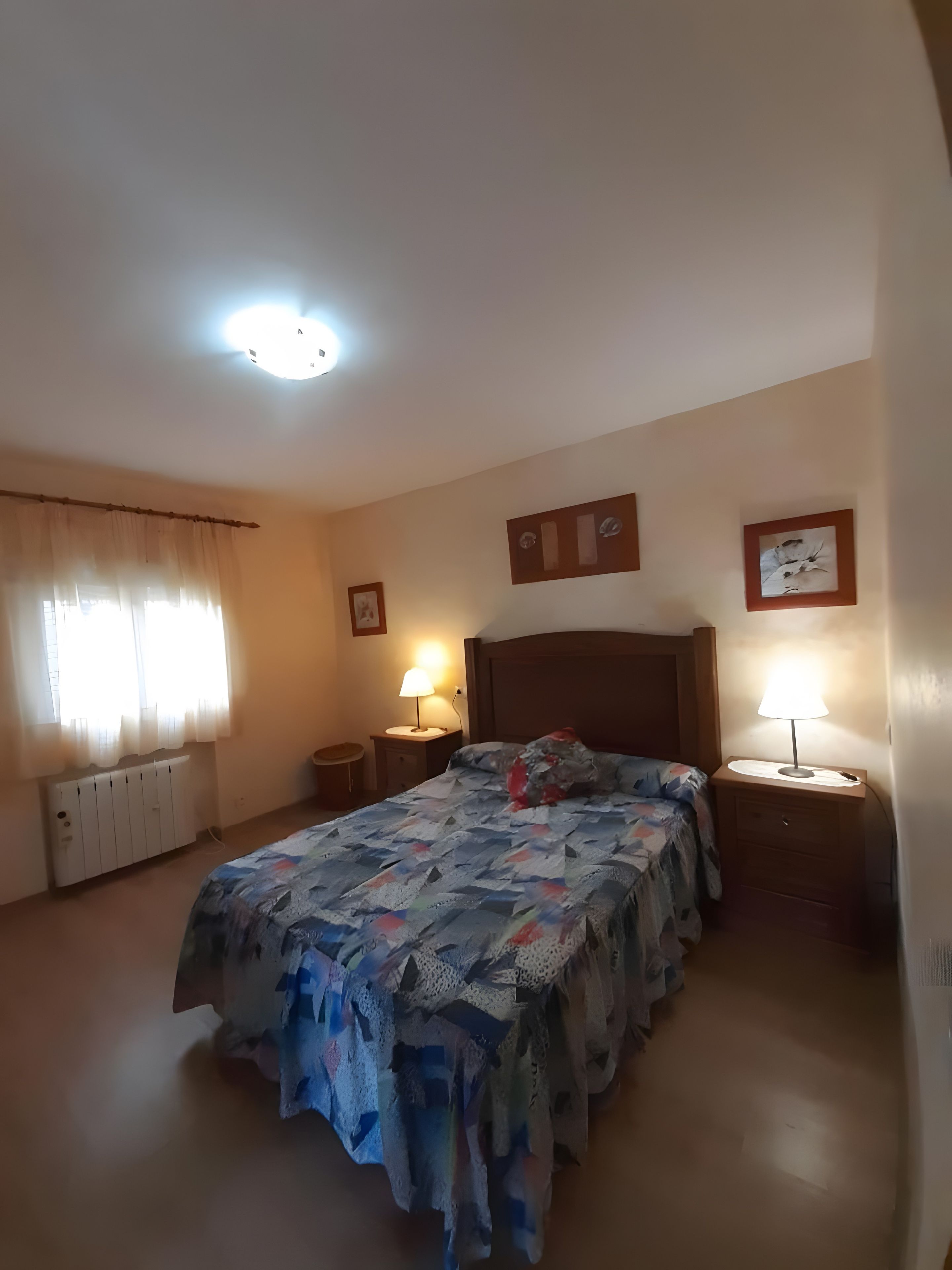 Un dormitorio con cama, dos mesitas de noche con lámparas, un radiador y una ventana con cortinas.