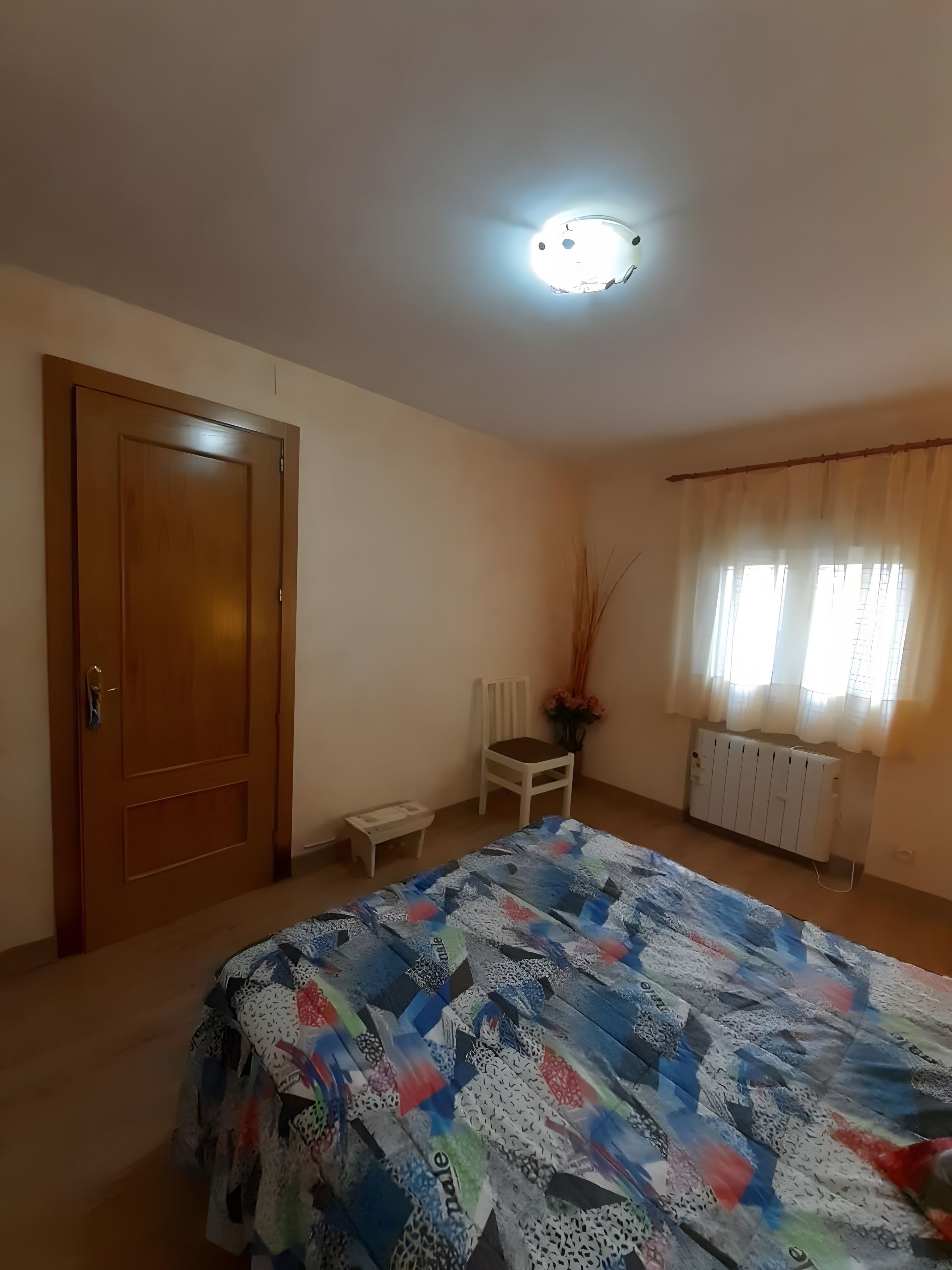 Un dormitorio con puerta de madera, una silla blanca, un radiador, una ventana con cortinas y una cama con edredón estampado.