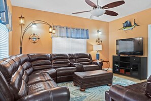 A living room with a sectional sofa, a ceiling fan, a flat-screen TV, and decorative wall art. - Seas the Day Retreat | GolfCart, LazyRiver & Beach (Horry County)