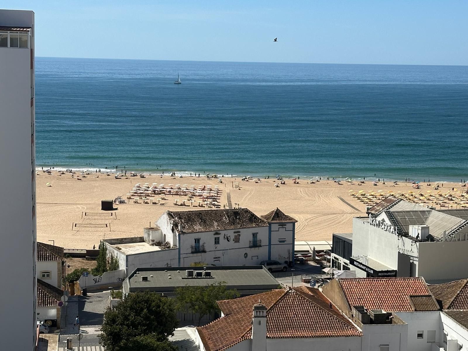 Blick auf die Küste mit Strand, Gebäuden und einem Segelboot auf dem Wasser.