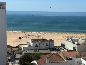 Vista costeira com praia, edifícios e um veleiro na água.
