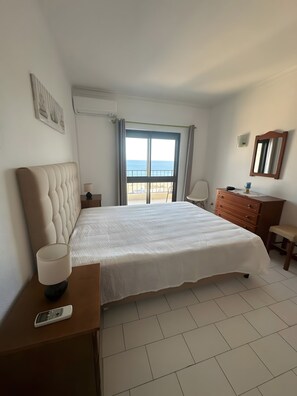 Ein Hotelzimmer mit einem großen Bett, einem Nachttisch mit Lampe, einem Schminktisch, einem Spiegel und Meerblick durch ein Fenster.