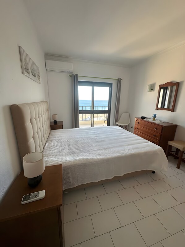 Quarto de hotel com uma cama grande, mesa de cabeceira com luminária, cômoda, espelho e vista para o mar pela janela.