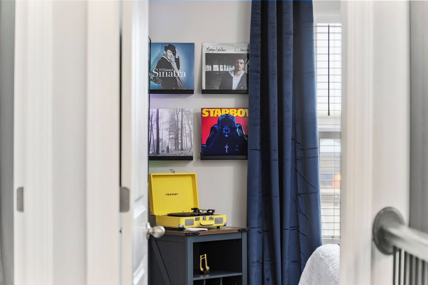 Ein Zimmer mit einem Bett, einem kleinen Tisch mit einem gelben Gegenstand und Wandkunst, darunter ein Poster mit dem Wort 'STARBOY'.