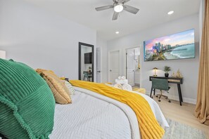 A hotel room with a bed, a desk, a chair, a ceiling fan, and a painting on the wall. - HGTV-Featured Beach House w/ Glow Arcade (Myrtle Beach)