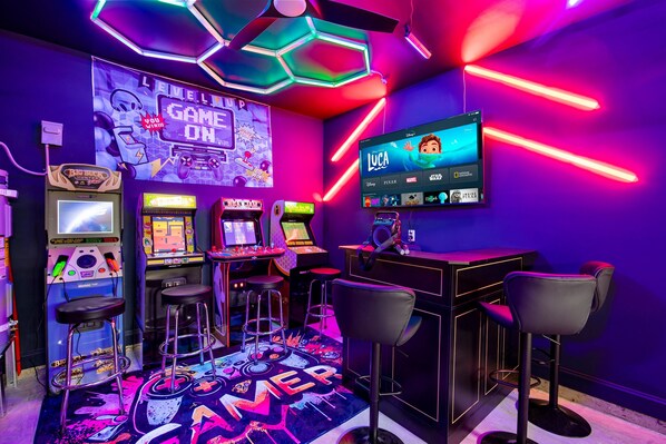 Zona de juegos con máquinas recreativas, una pantalla grande mostrando una película y taburetes de bar.