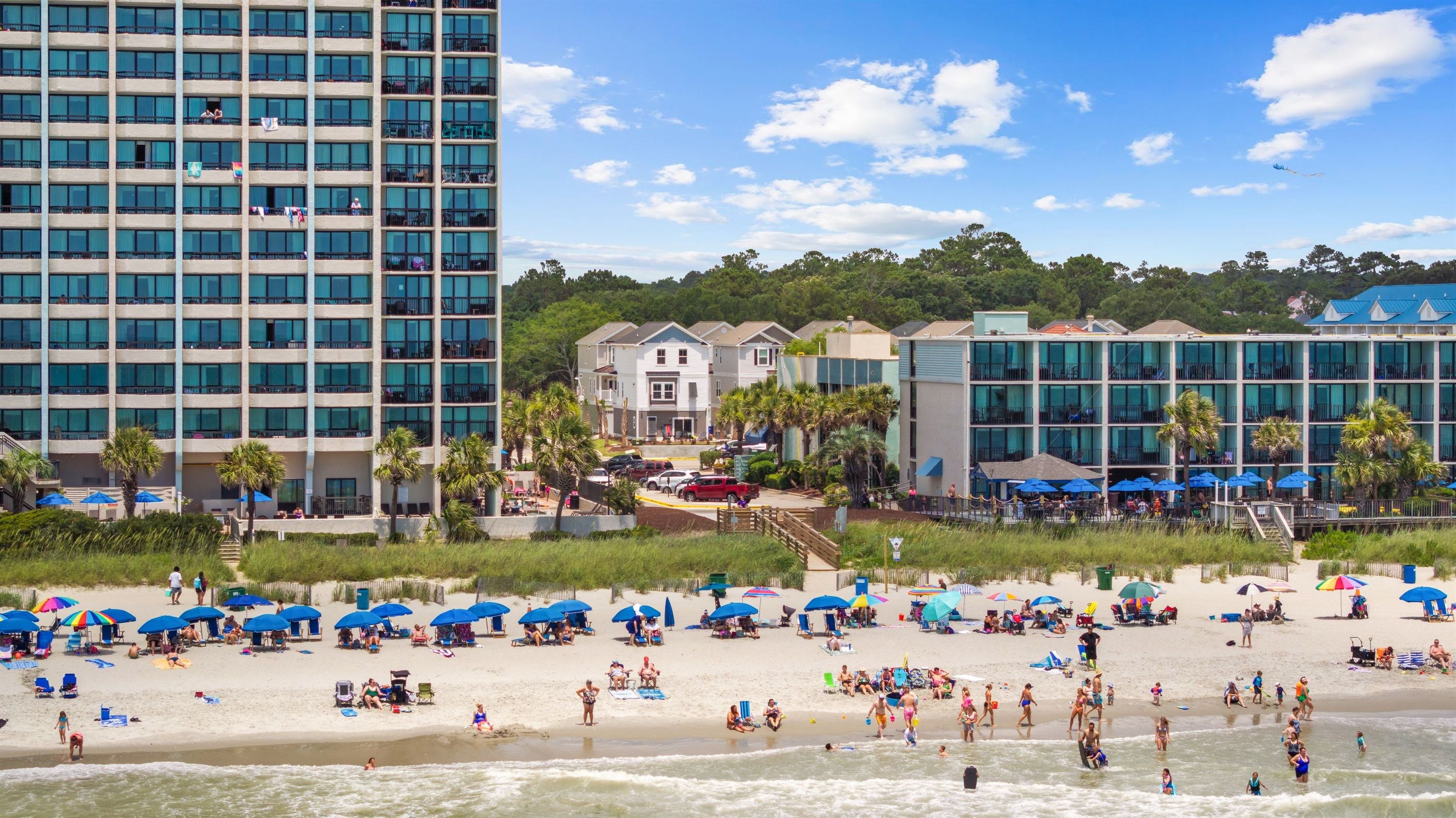 A beach with umbrellas and people, a hotel building, and a parking area.