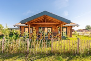 A wooden cabin with a covered porch, surrounded by a wooden fence and greenery. - Ferienhaus mit Eigener Sauna und Schoener Terrasse (Toenning)