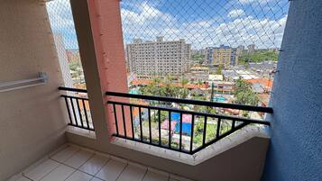 Uma varanda com grade de arame, piso de azulejos e vista para edifícios e vegetação ao redor.