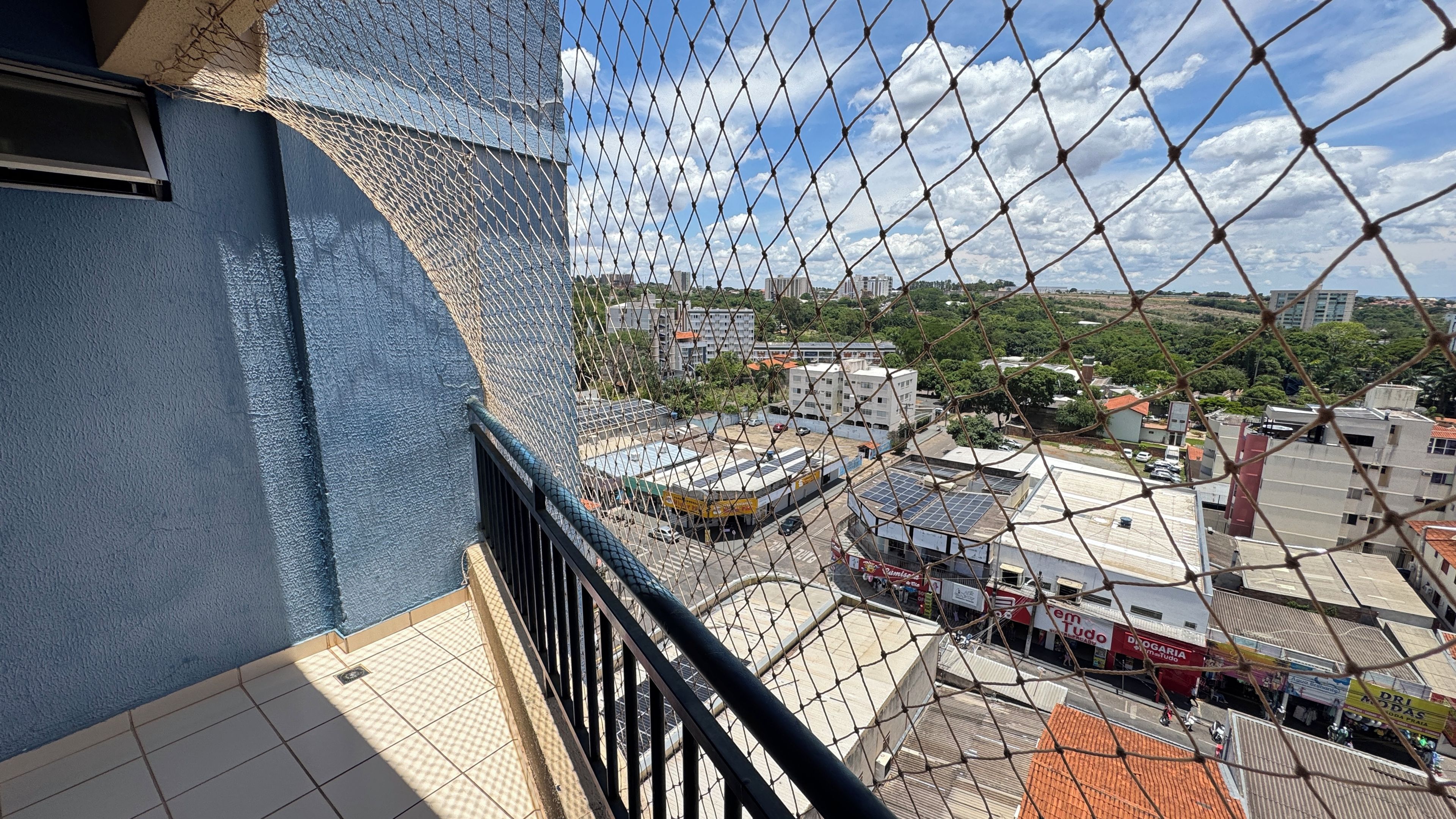 Um edifício alto com fachada azul, uma varanda com guarda-corrimão preto e vista para um horizonte urbano com diversos edifícios e céu claro.