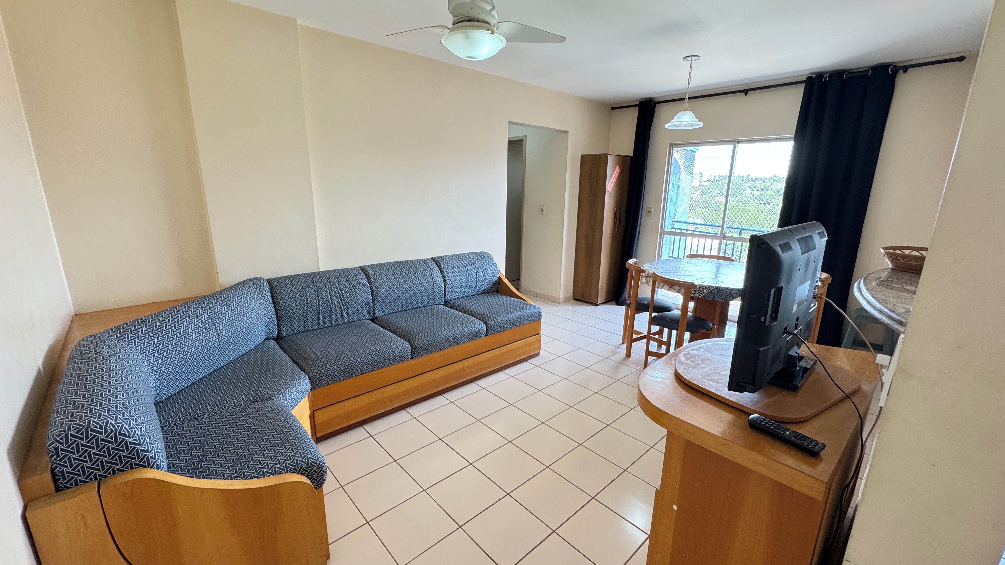 Uma sala de estar com sofá, televisão sobre um suporte de madeira, mesa de jantar com cadeiras e uma varanda com vista.