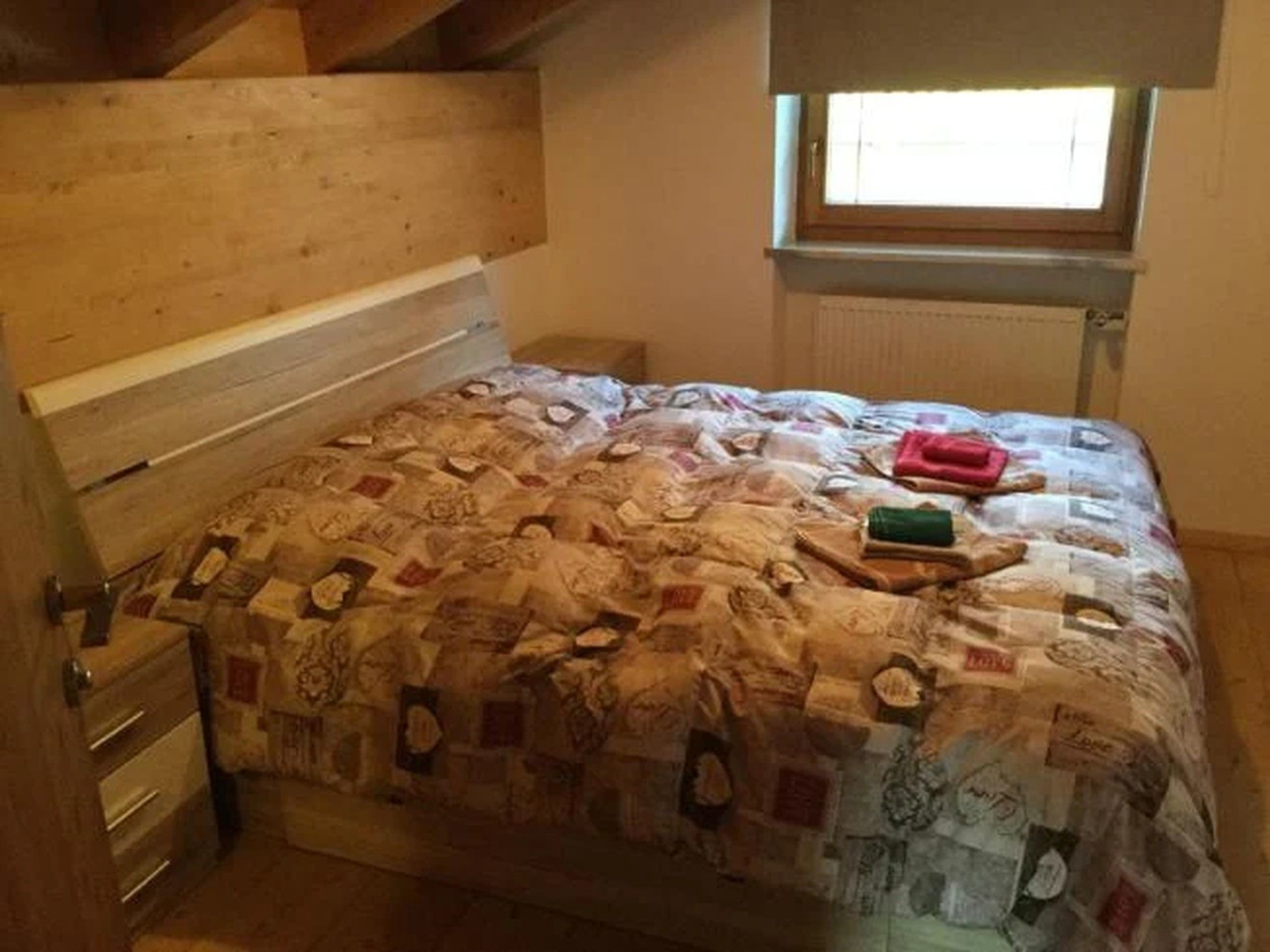 A bedroom with a wooden headboard, a quilted bedspread, a window with blinds, and a radiator.