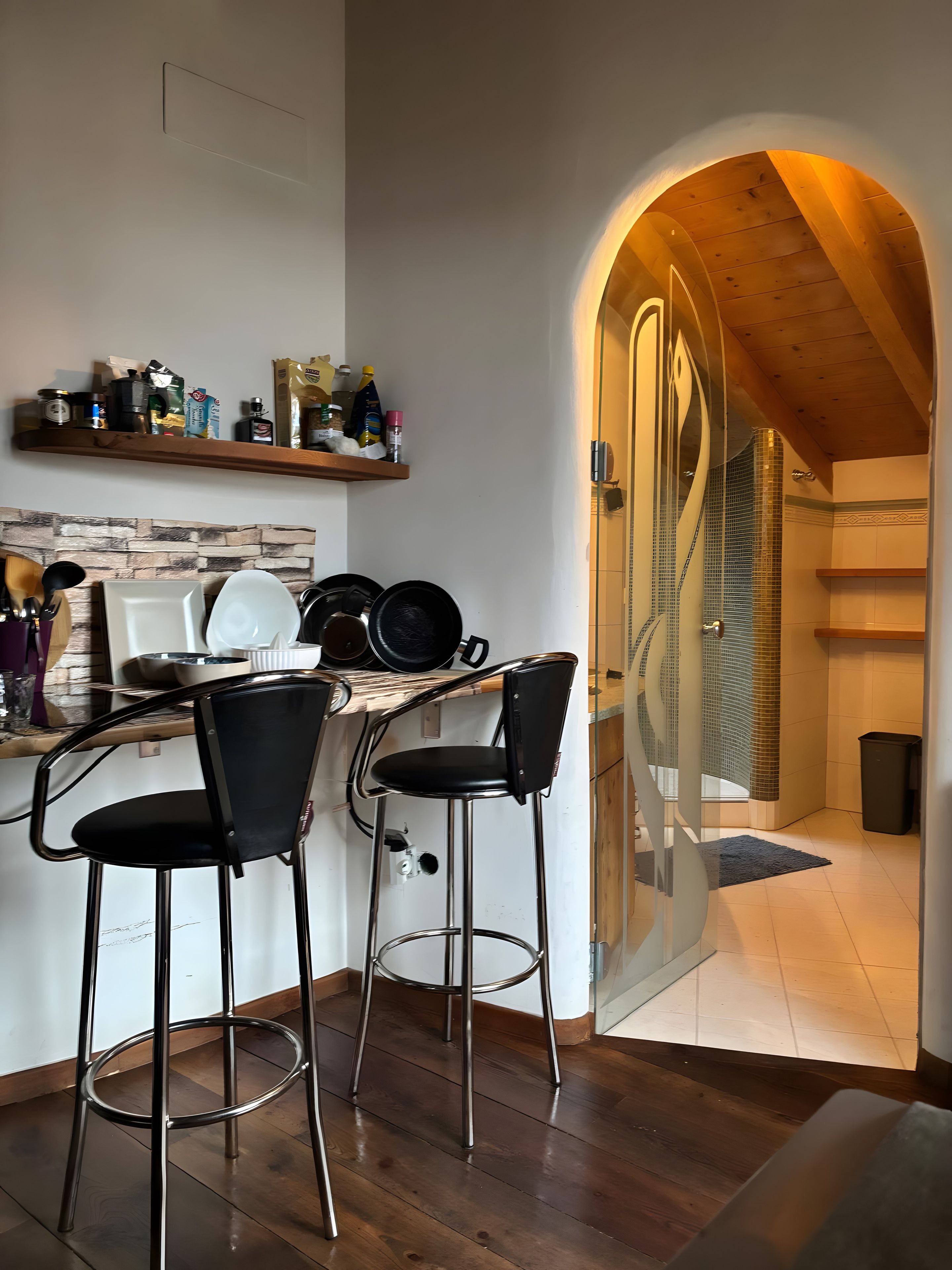 A kitchen with two bar stools, a wooden ceiling, and a glass door leading to a shower area.