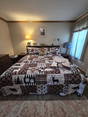 A bedroom with a large bed covered in a patterned quilt, a nightstand with a lamp, a framed picture on the wall, and a window with curtains.