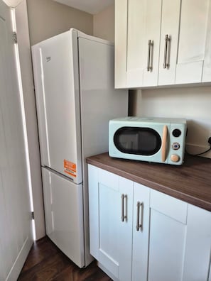 Cozinha compacta com geladeira branca, micro-ondas na bancada e armários brancos com puxadores prateados.