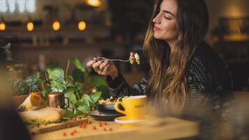 A person dining at a table with a cup and a plate of food, surrounded by plants and a window.