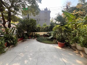 A paved pathway flanked by potted plants and trees, leading to a building with a grey facade.