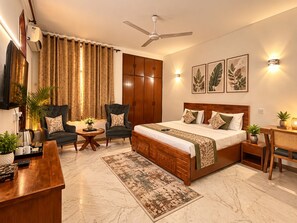 A hotel room with a bed, two armchairs, a coffee table, a desk, a TV, a ceiling fan, and wall-mounted lights.
