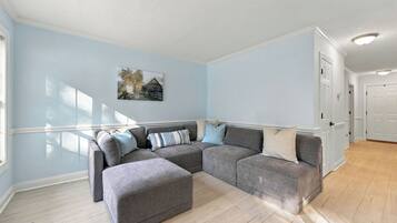 Una habitació amb un sofà seccional gris, un otomà petit, parets de blau clar, un quadre a la paret i una porta al fons.