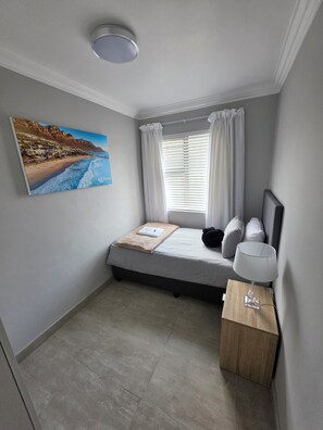 A bedroom with a bed, a bedside lamp, a window with curtains, a painting on the wall, and a small wooden side table. - Perfect 2-bedroom apartment in a delightful Cape Town neighbourhood (Cape Town)